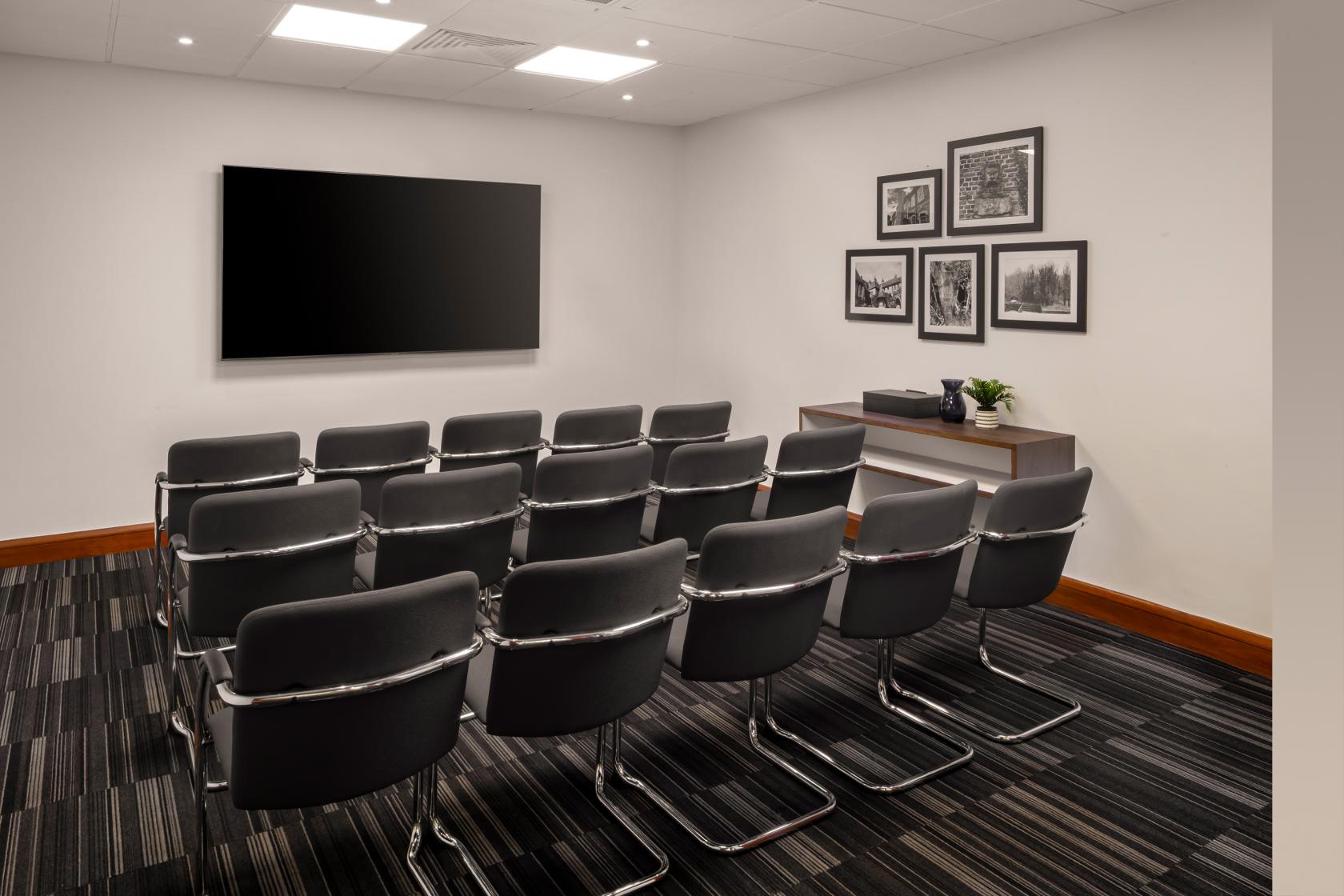 A boardroom with rows of black chairs, a large wall-mounted screen, and framed photos at Holiday Inn London Bexley.