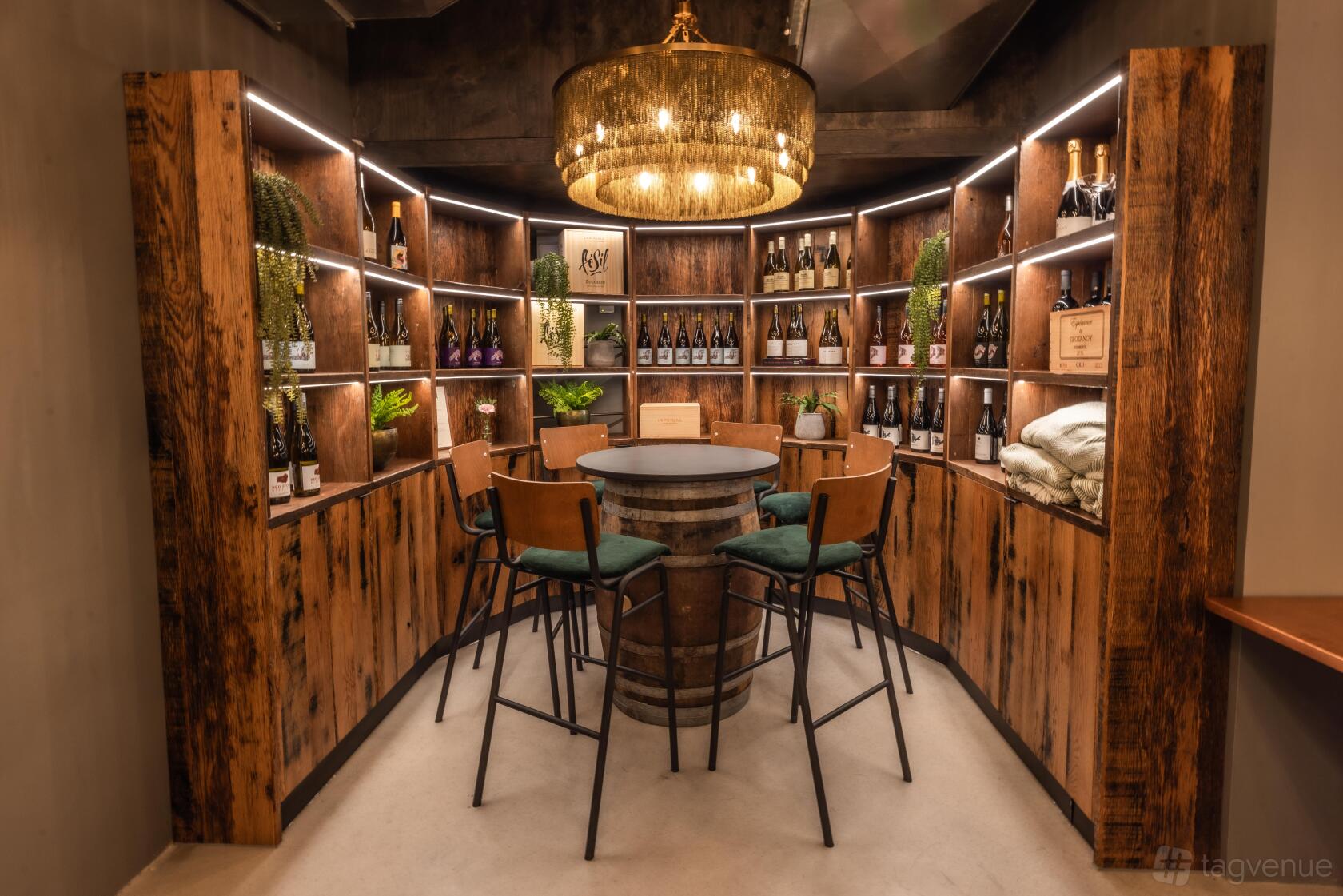 An event space in a bar with wood-paneled wine shelves, a round barrel table, and a chandelier at Vagabond Battersea.