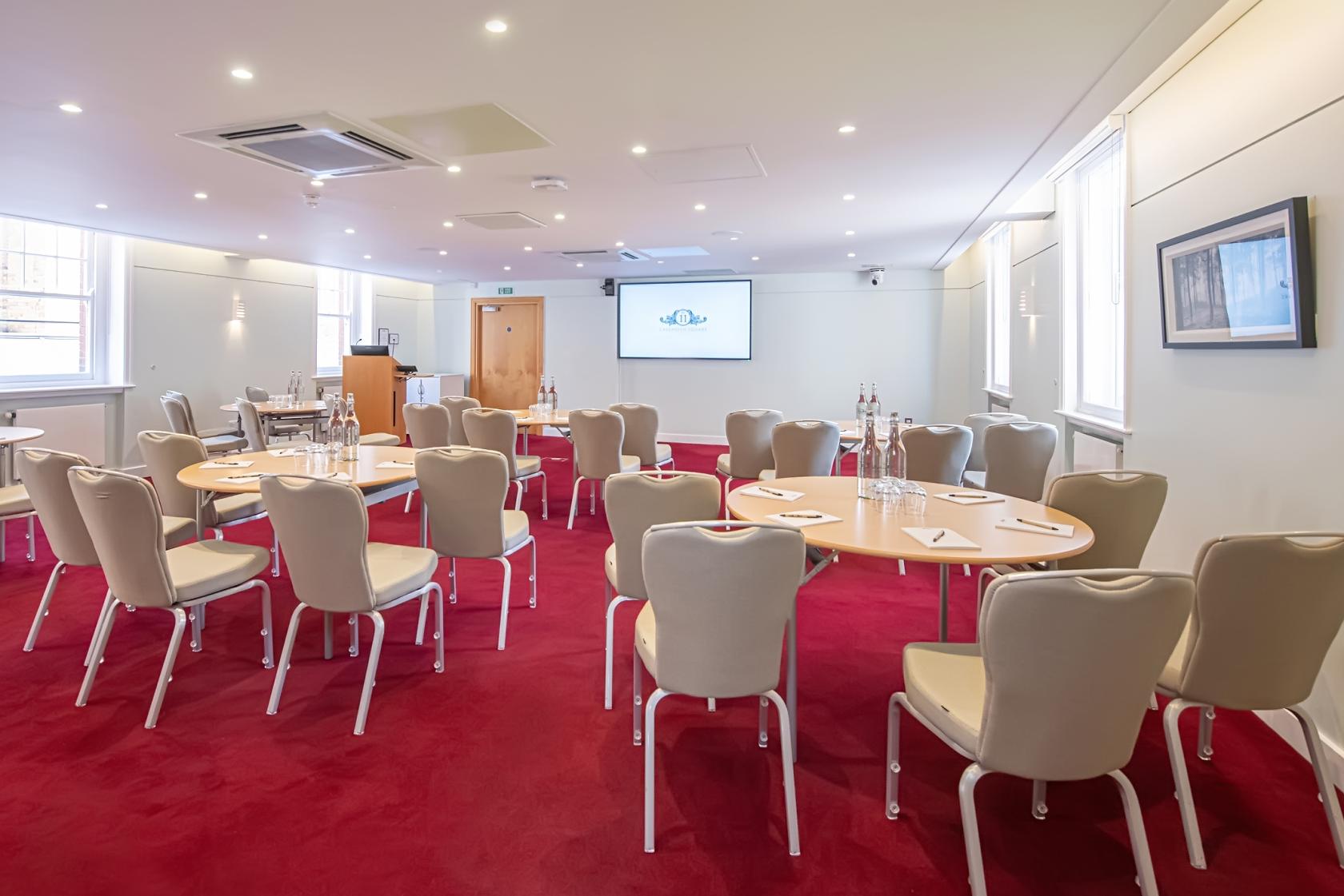 A function room with round tables, cream chairs, red carpeting, and a wall-mounted screen at No.11 Cavendish Square.