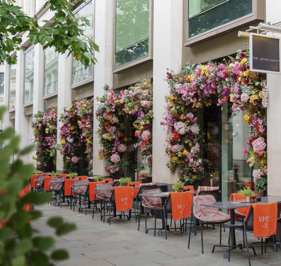 A restaurant patio with sidewalk seating, orange chairs, and large floral displays at The Anthologist.