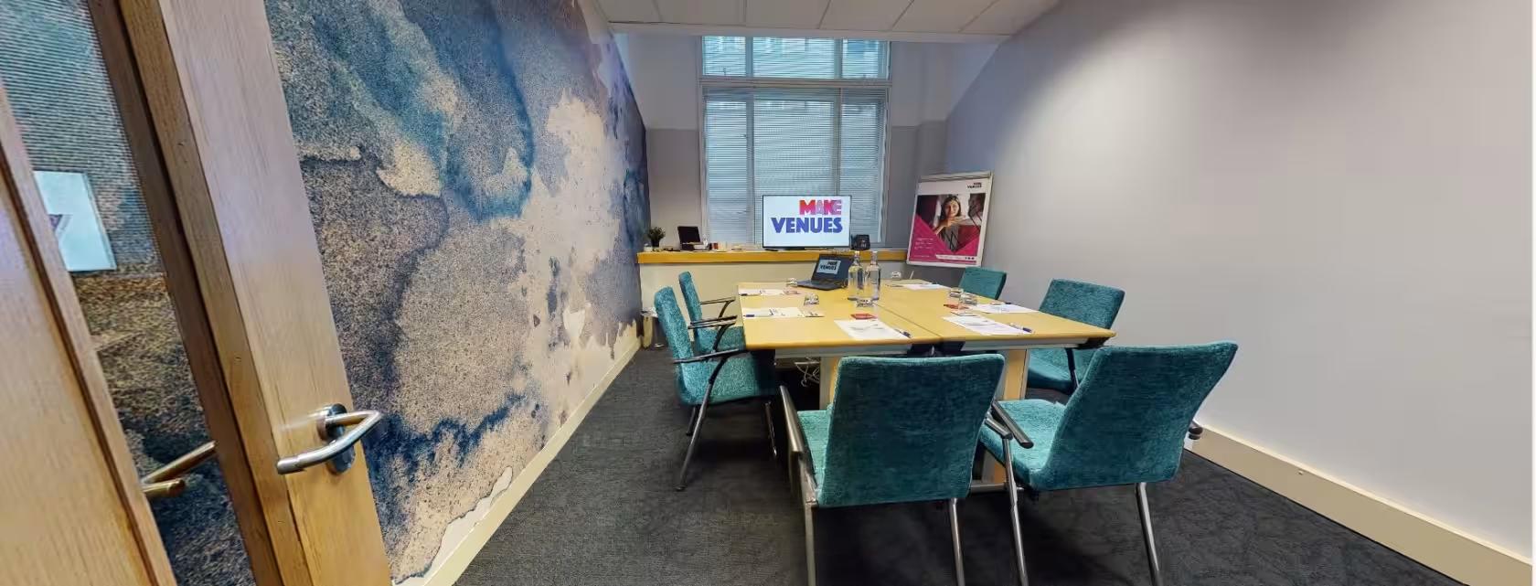 A meeting room with a textured accent wall, large window, and rectangular table with teal chairs at Broadway House.