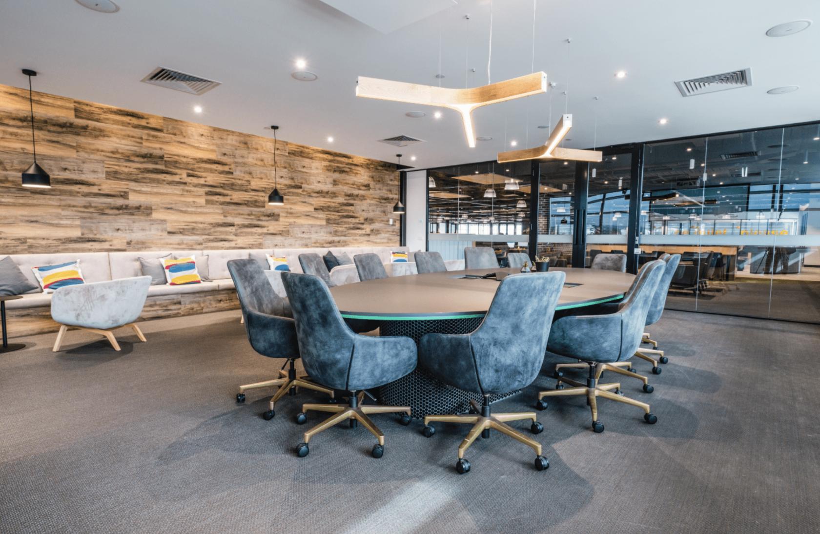 A boardroom with a large oval table, velvet swivel chairs, wood accent wall, and built-in sofa at Waterman at Caribbean Park.