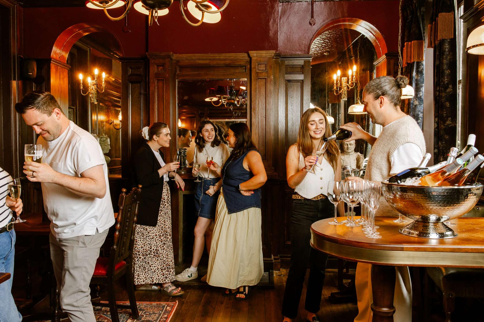 A dining room with dark wood paneling, chandeliers, and guests mingling near a bar at Bow Street Tavern.