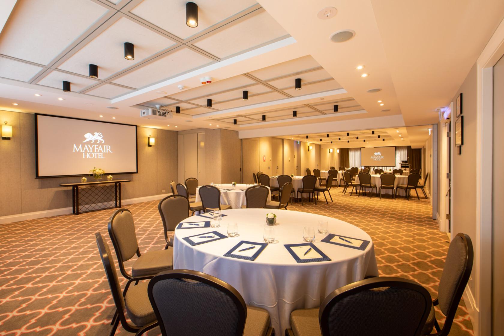 A function room with round tables, banquet chairs, a large projector screen, and patterned carpet at Mayfair Hotel Adelaide.