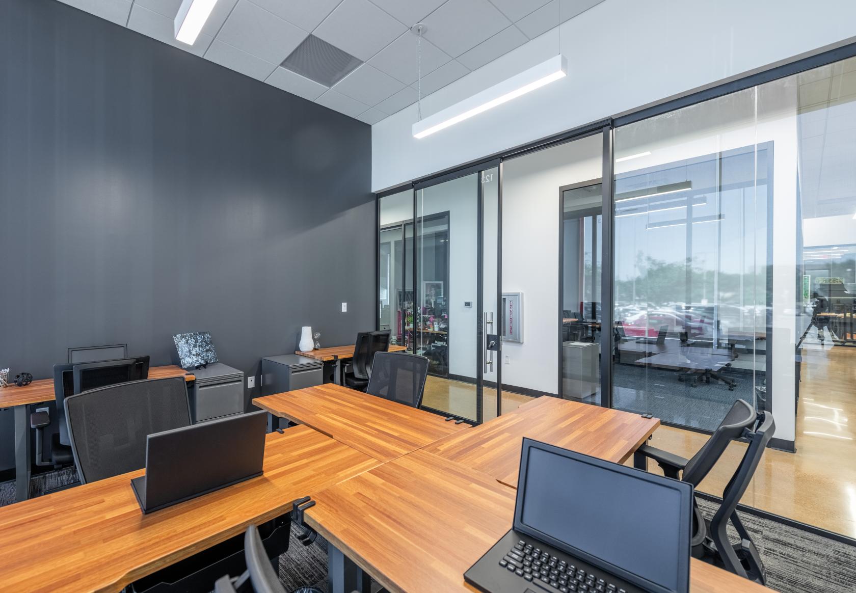 A meeting room with wood tables, black chairs, laptops, and glass walls at Venture X Dallas – Braniff Centre.