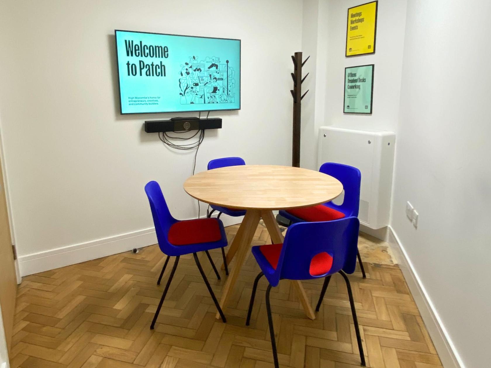 A meeting room with a round wooden table, blue chairs, a wall-mounted screen, and a coat rack at Patch High Wycombe.