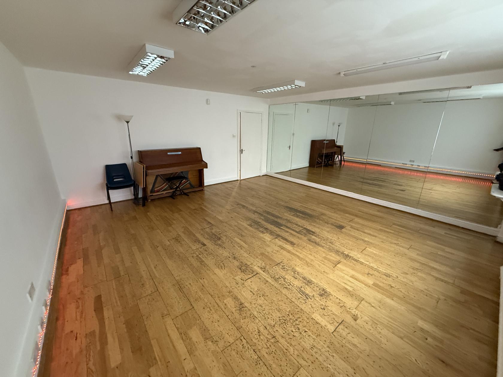A rehearsal room with wood flooring, a wall of mirrors, and an upright piano at artsHouse.