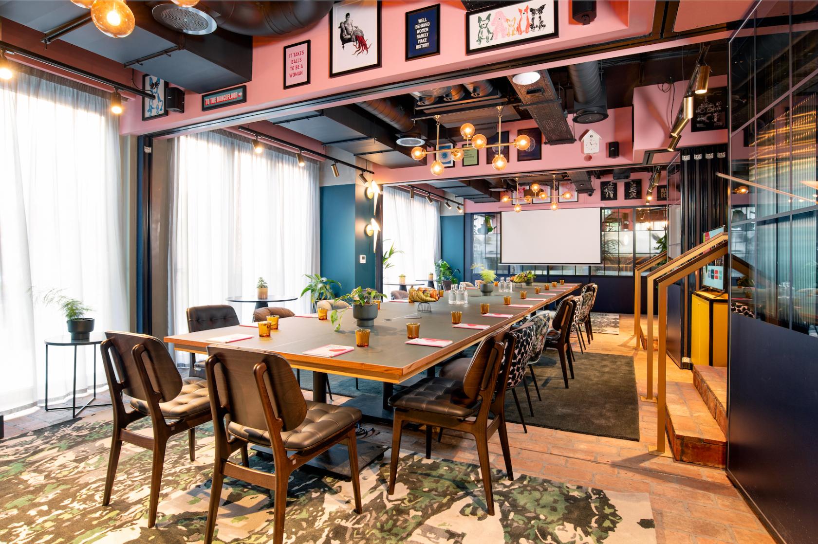 An event space with floor-to-ceiling windows, long banquet tables, potted plants, and eclectic wall art at Yotel Manchester Deansgate.