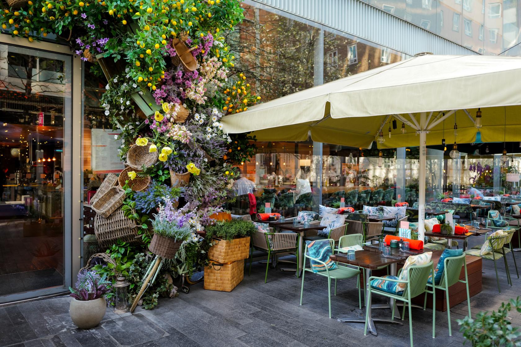 An outdoor restaurant terrace with umbrella-shaded tables, colorful cushions, and a floral display at The Refinery Bankside.