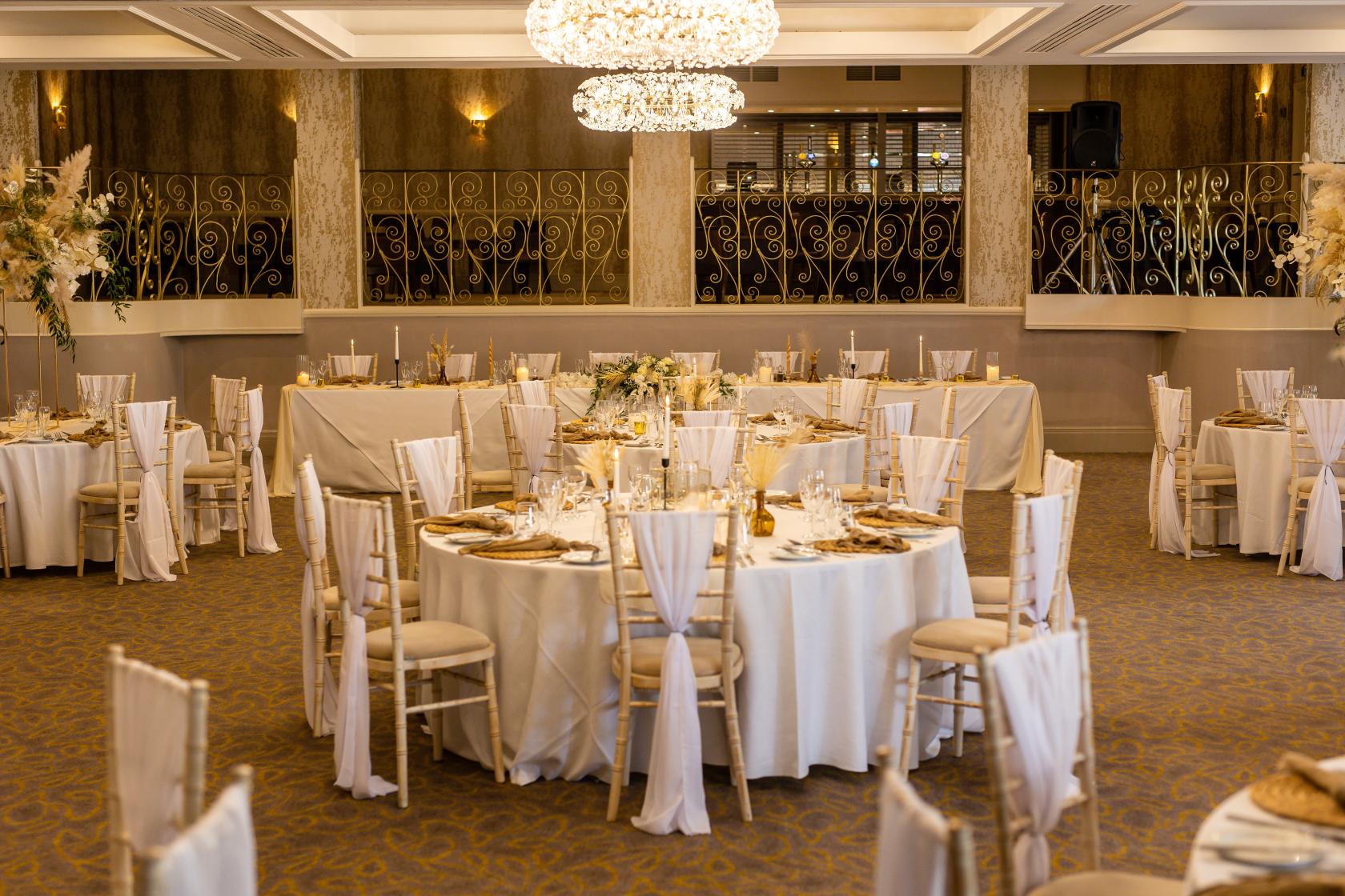 An event space in a hotel with round tables draped in white linens, gold plates, and chandeliers at Albrighton Hall Hotel.
