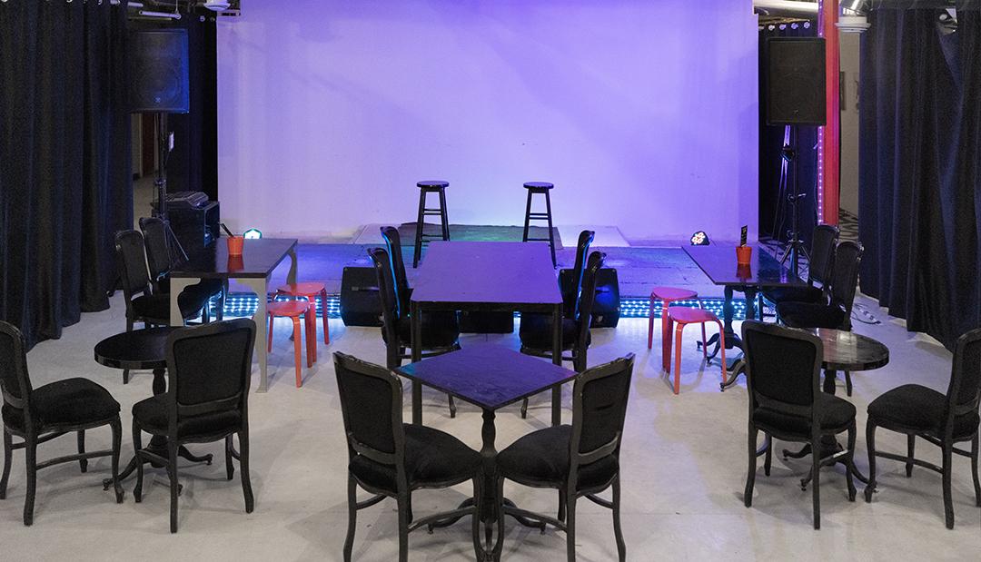 An event space with a small stage, black chairs, tables, and colored accent lighting at Primal Note Studios.