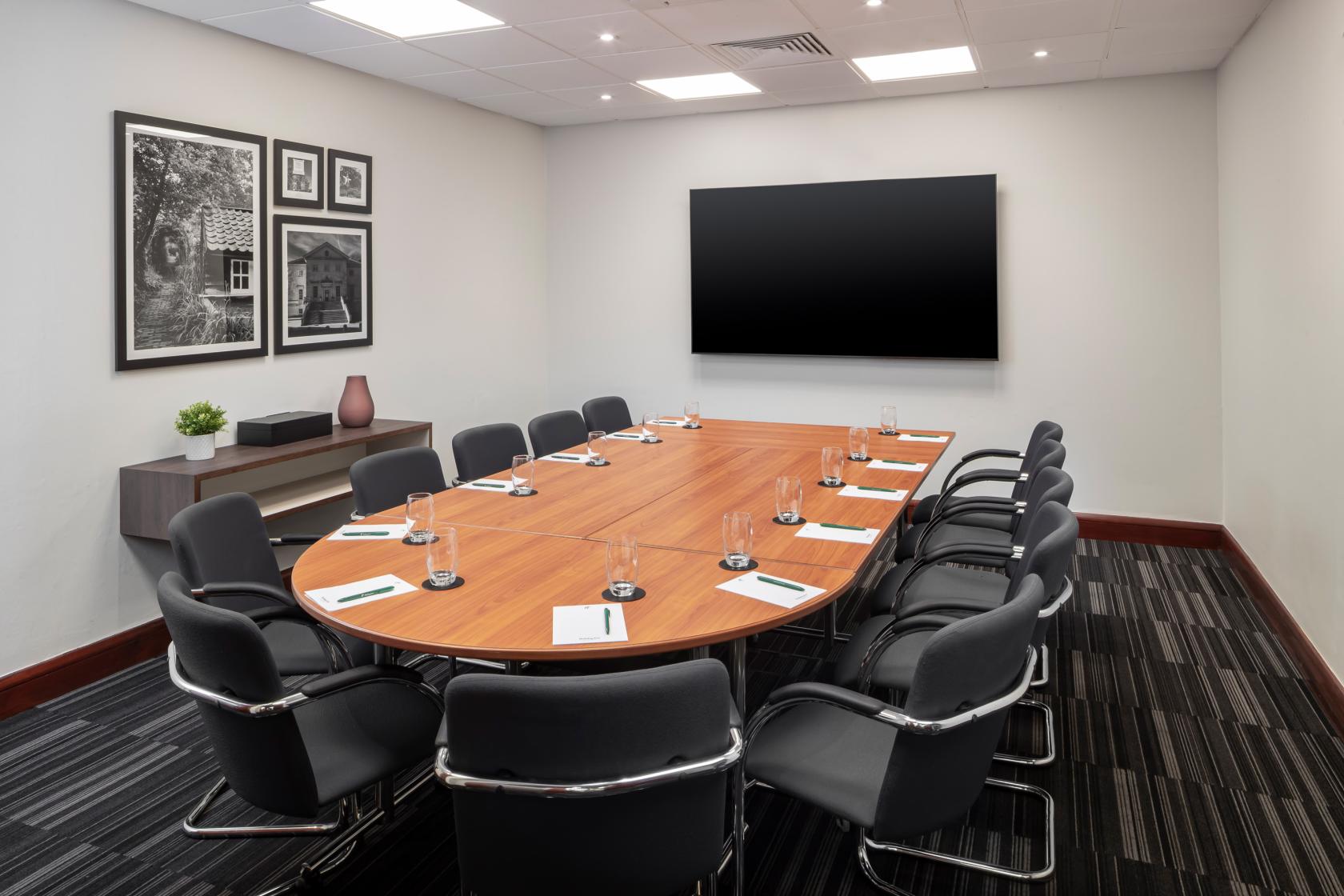 A boardroom with a large wooden conference table, black chairs, and a wall-mounted screen at Holiday Inn London Bexley.