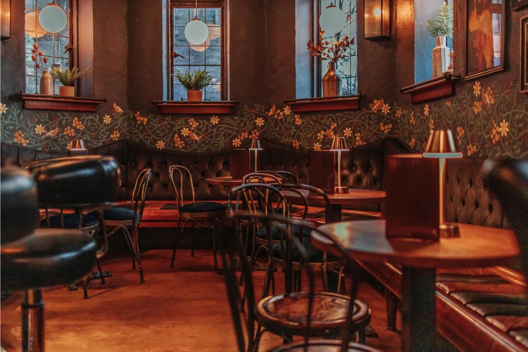 A bar in a pub with dark tufted booths, floral wallpaper, wooden tables, and warm ambient lighting at Òran Mór.