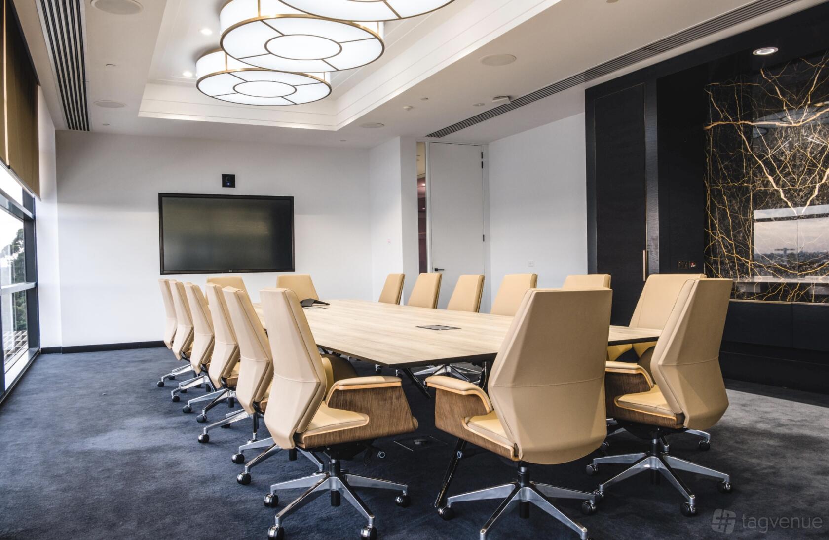 A boardroom with a long wooden table, tan swivel chairs, a large wall-mounted screen, and ring ceiling lights at Waterman at Chadstone.