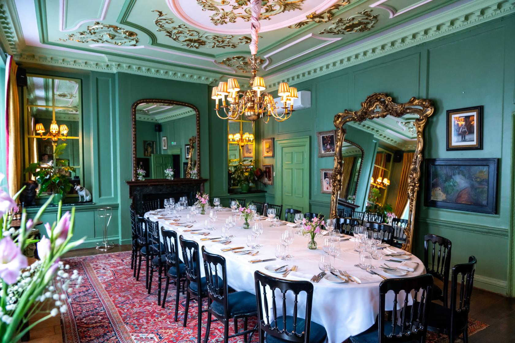 A dining room with ornate green walls, gilded mirrors, chandeliers, and an oval table with white linens at L'Escargot.