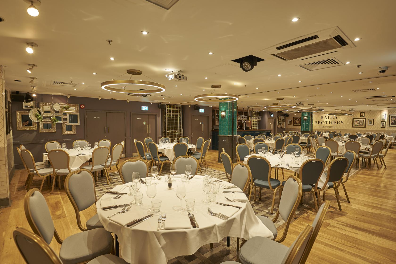 A function room with round tables dressed in white linens, cushioned chairs, and modern lighting at Balls Brothers Minster Court.