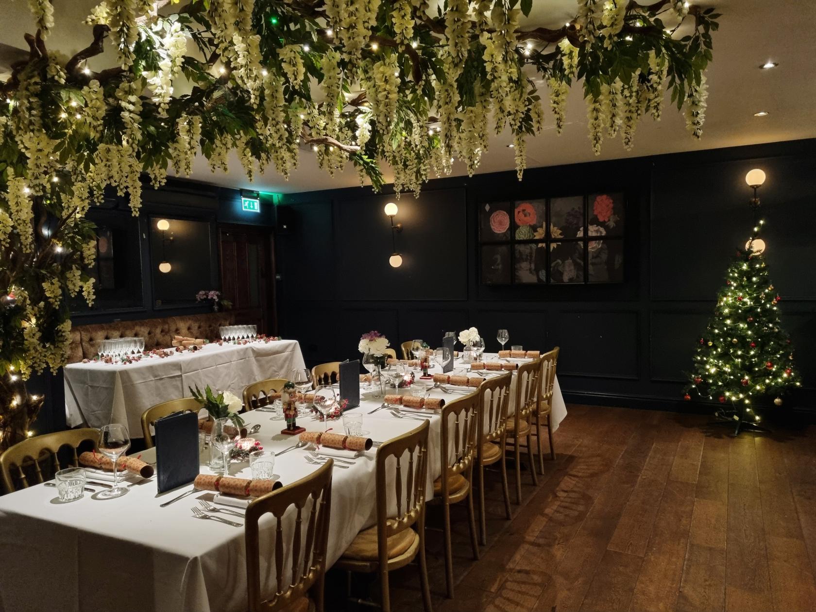 A dining room with long tables set with white linens, floral centerpieces, hanging greenery, and fairy lights at The Lost & Found Bristol.