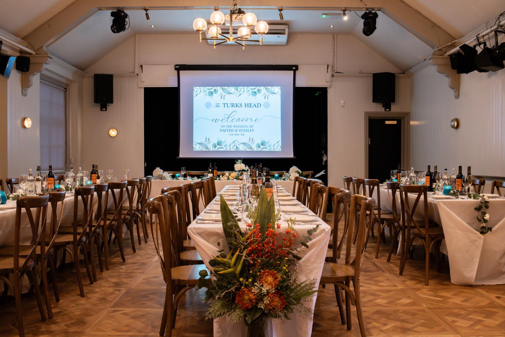 A function room with banquet tables, wooden chairs, white tablecloths, and a projector screen at The Turks Head.