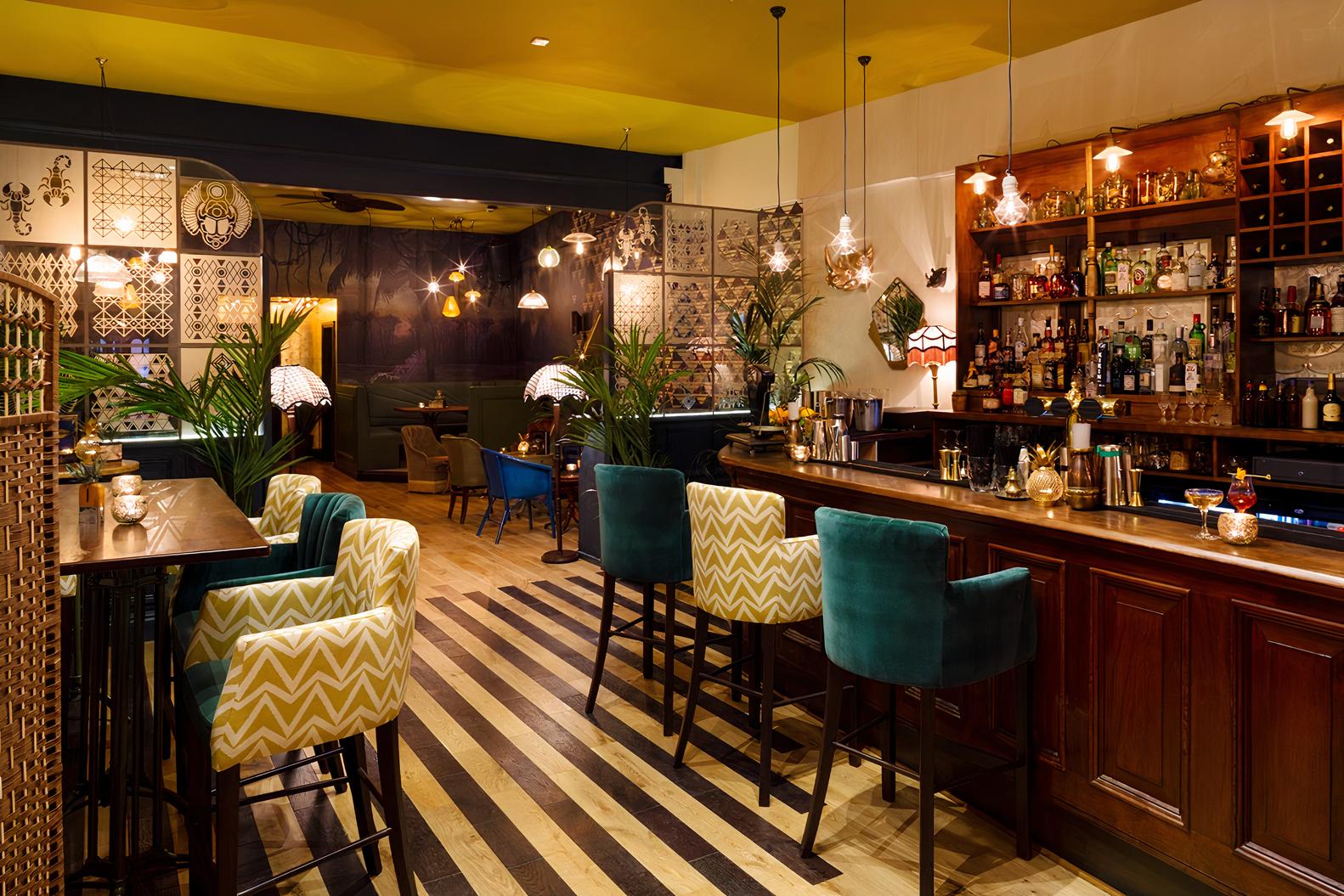 A restaurant with patterned armchairs, wooden bar, hanging pendant lights, and eclectic decor at Hoxley and Porter.