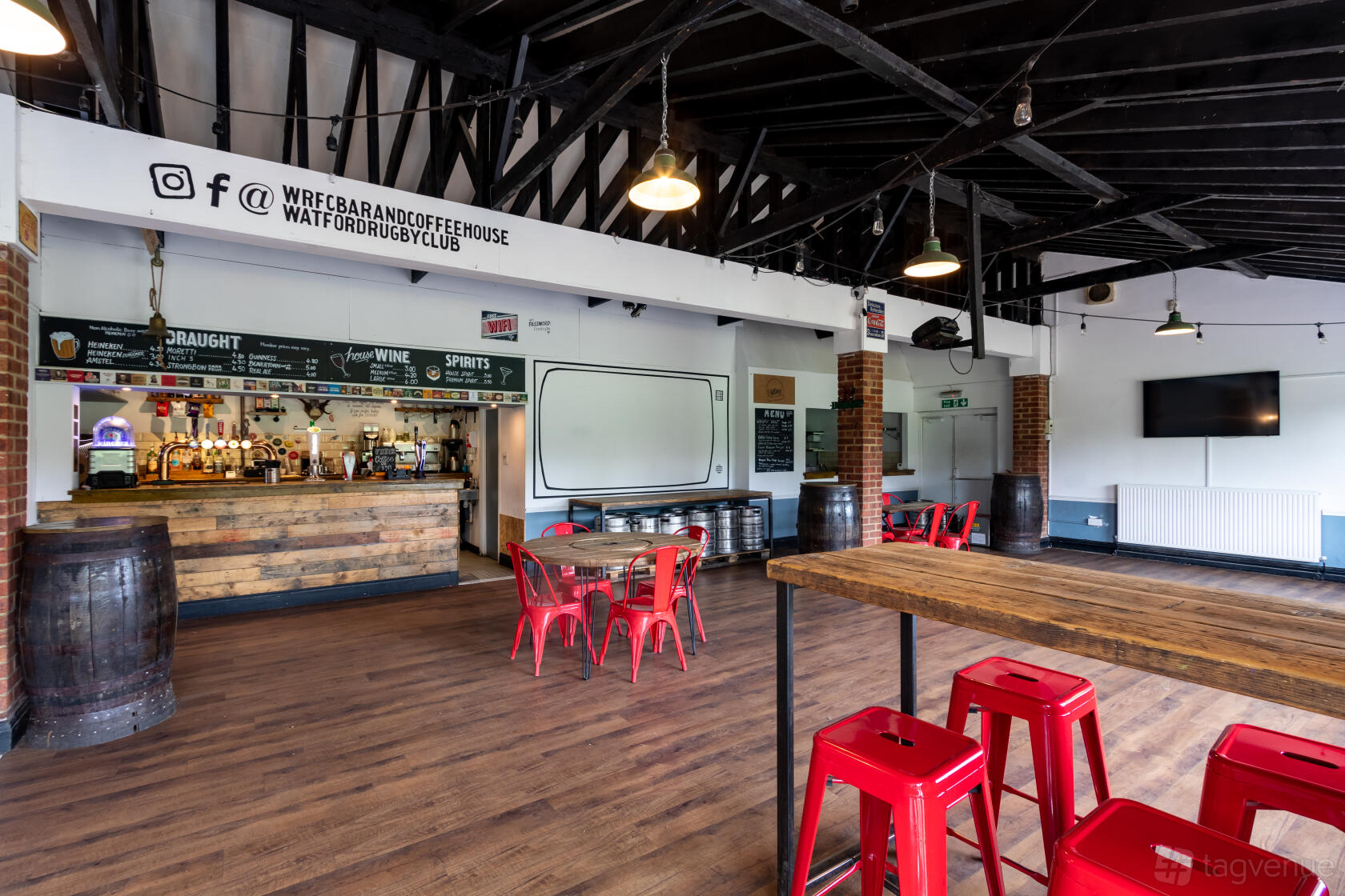 A bar with exposed beam ceiling, high wooden tables, red metal stools, and a large bar counter at WRFC BAR & COFFEE HOUSE.