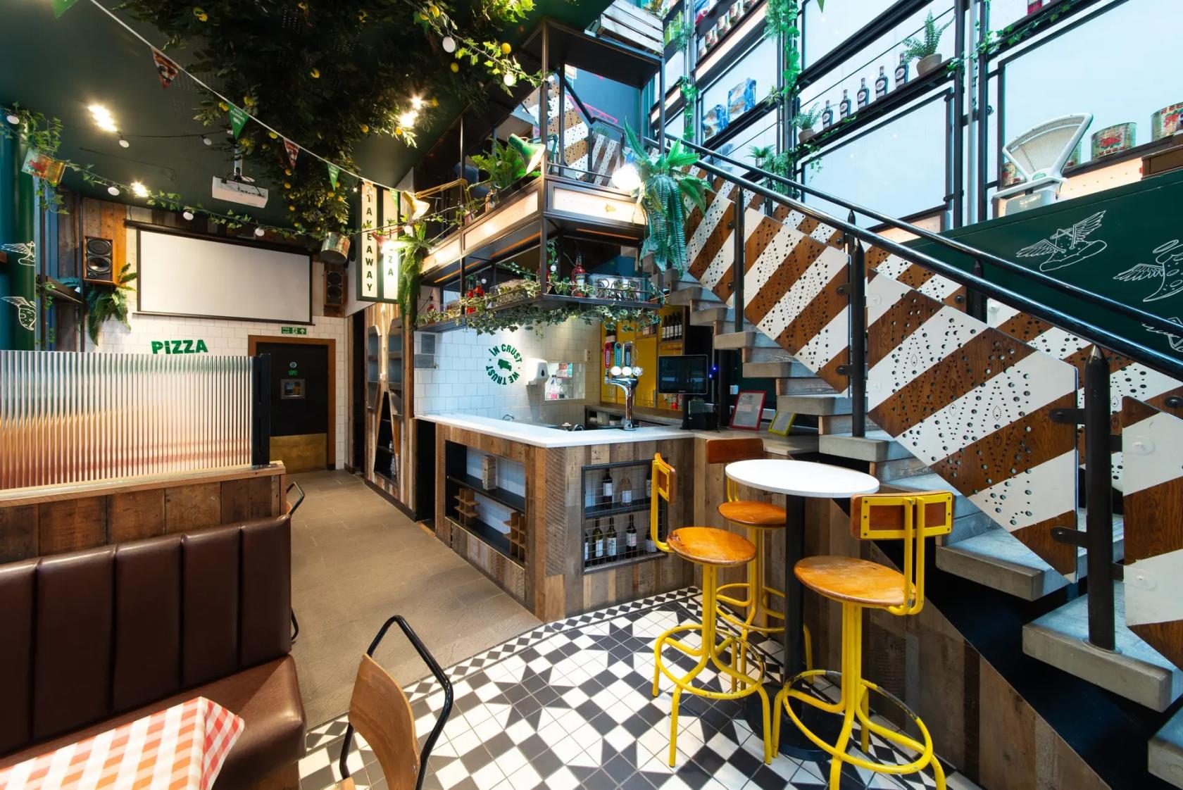 A bar in a restaurant with yellow bar stools, round tables, and an industrial staircase at Pizza Pilgrims Liverpool Street.