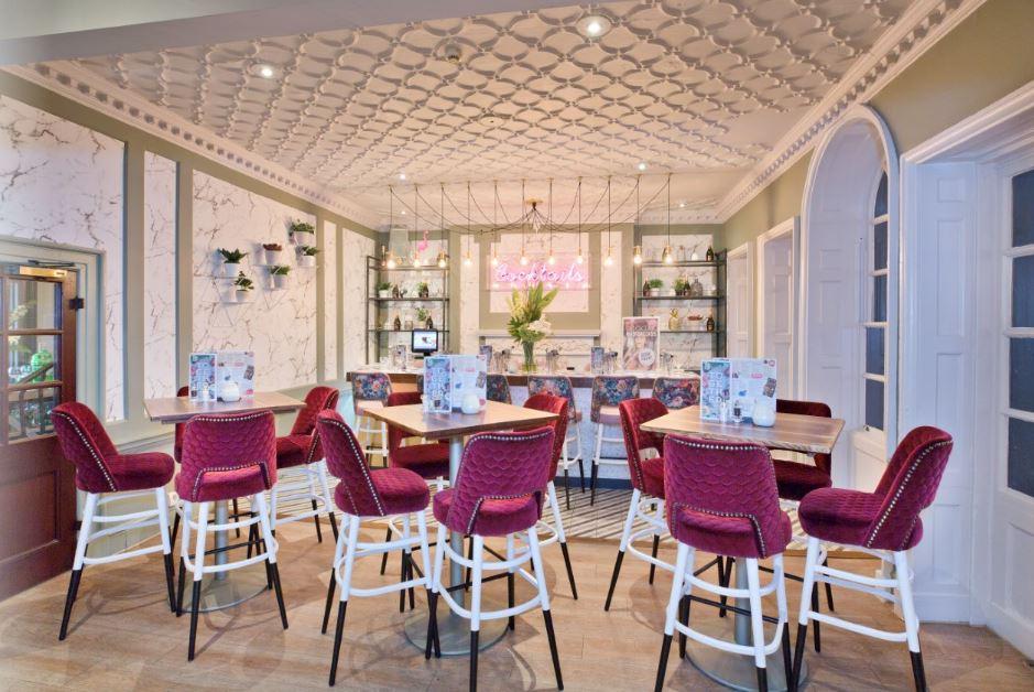 A bar with high tables, red velvet bar stools, a decorative ceiling, and a neon sign behind the counter at Slug and Lettuce Bath.