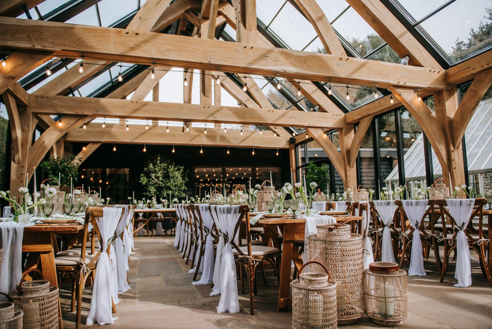An event space in a barn with exposed timber beams, glass ceiling, and tables draped with white linens at Chapel House Estate.