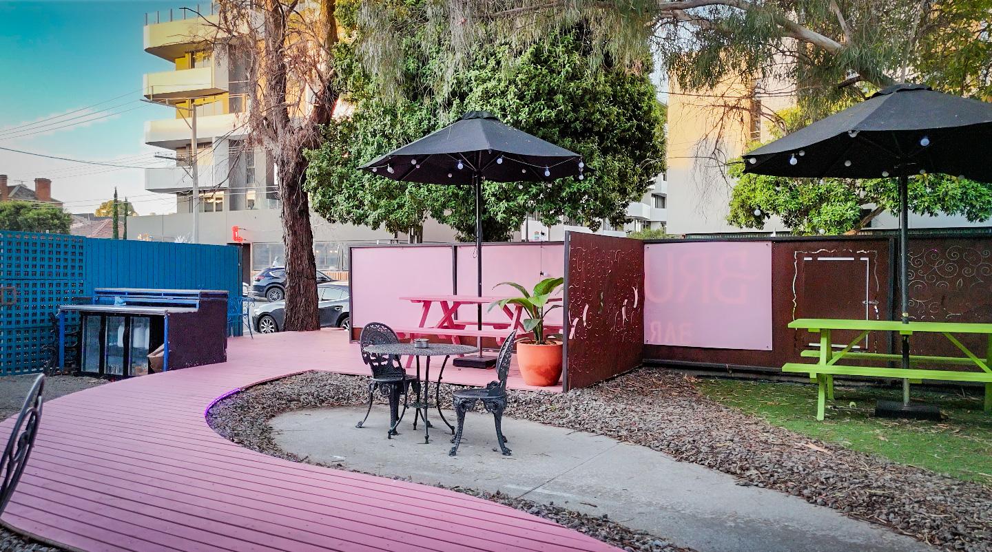 An outdoor pub area with black metal chairs, pink picnic tables, and umbrellas at Aces Bar @ Brunswick Aces.