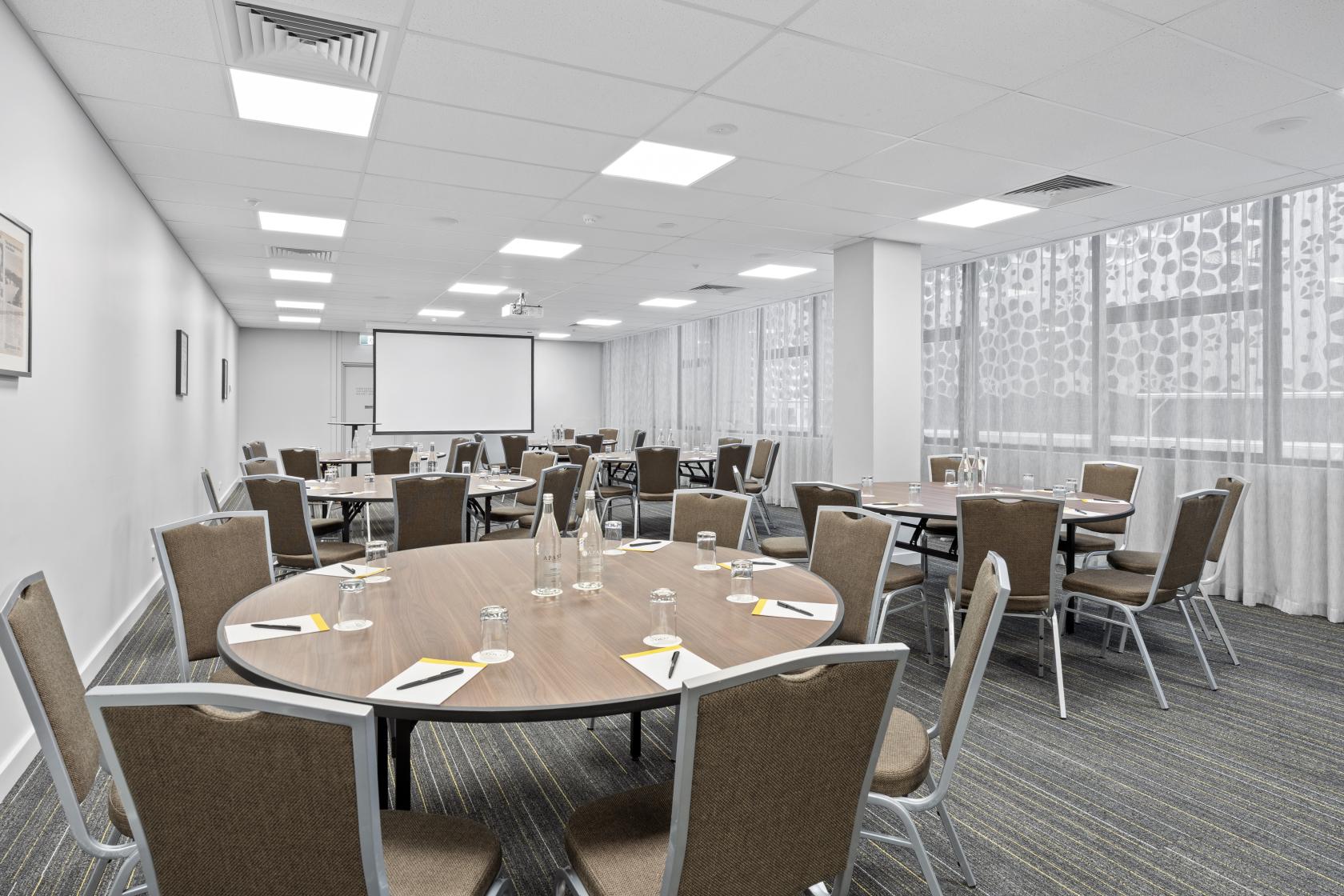 An event space with round tables, brown chairs, and floor-to-ceiling windows at Voco Hotel Brisbane City Centre.