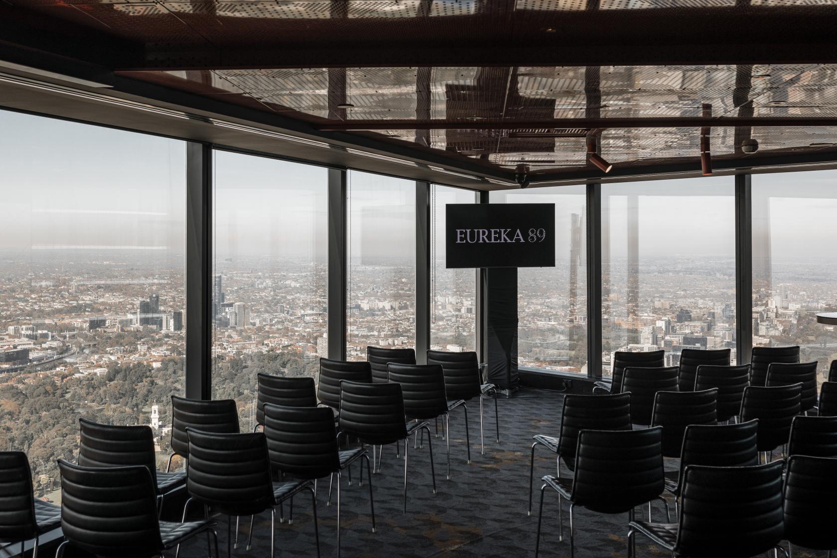 A function room in a restaurant with floor-to-ceiling windows, city views, and rows of black chairs at Eureka 89.