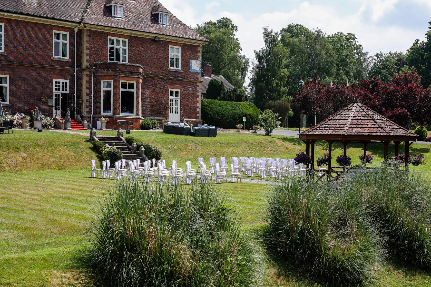 An outdoor hotel space with a lakeside pavilion, white chairs on a lawn, and landscaped gardens at Albrighton Hall Hotel.