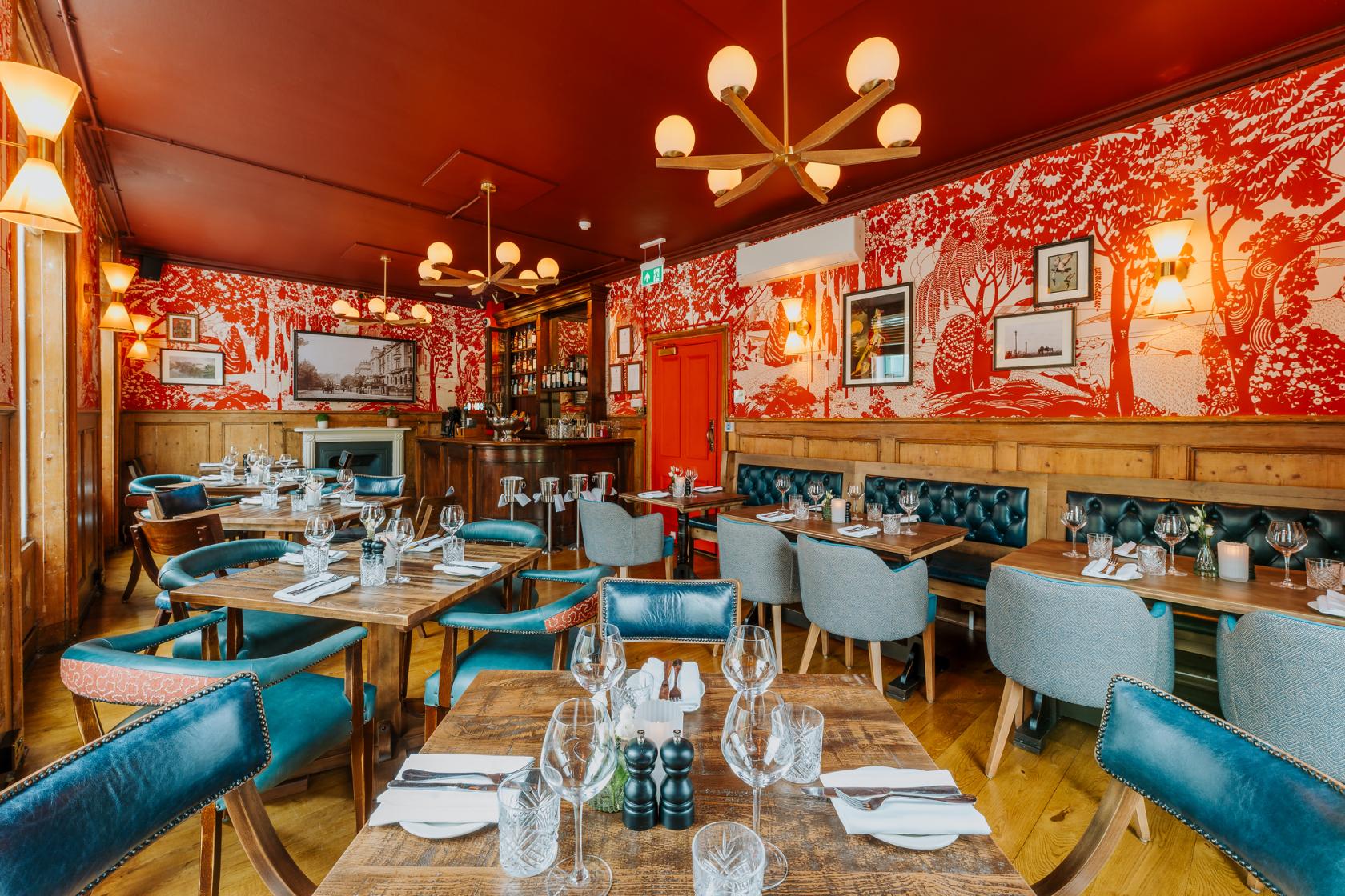 A pub dining area with red patterned wallpaper, tufted leather seating, and round chandeliers at The Engineer.