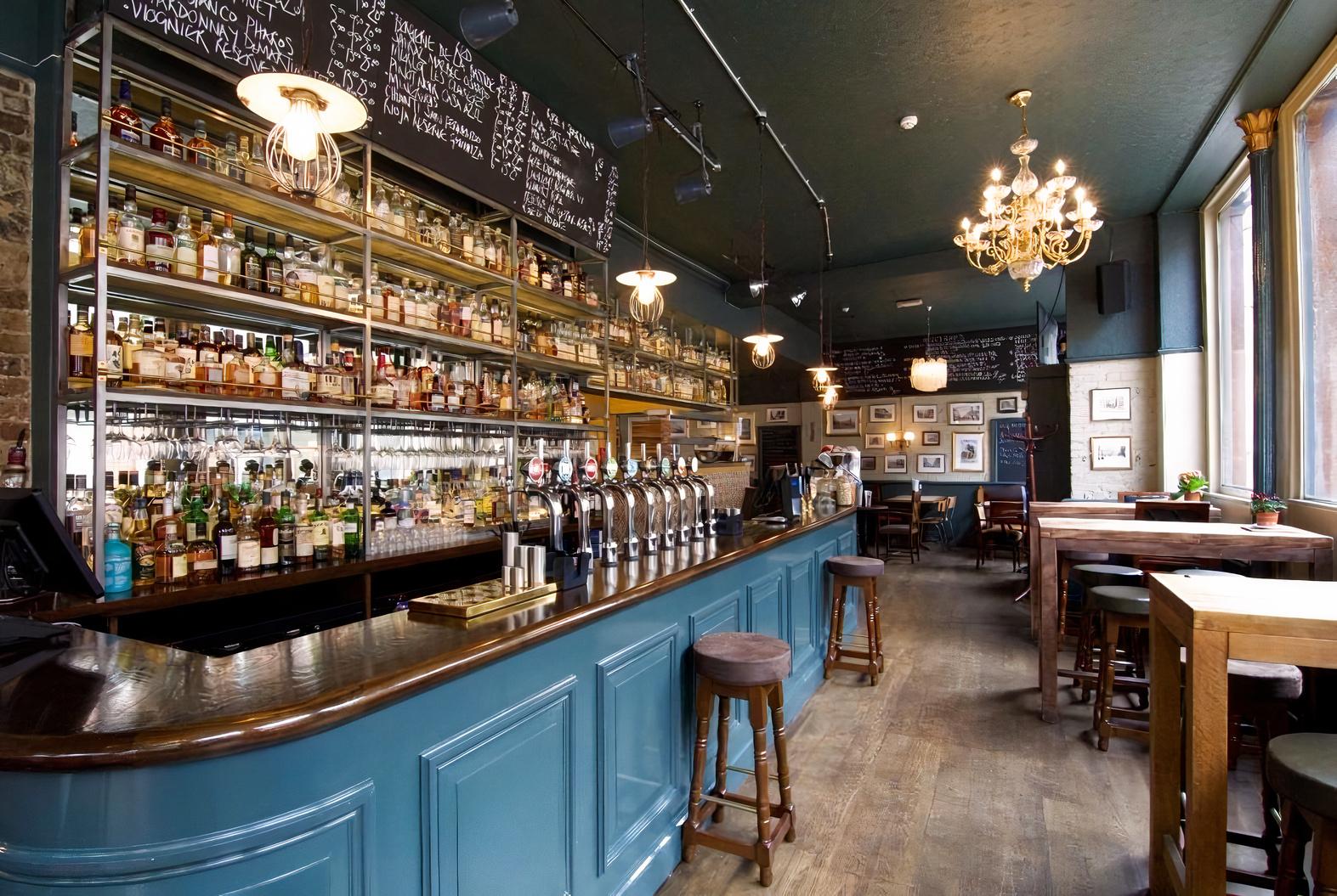 A pub with a long blue bar, wall-mounted shelves of liquor bottles, chalkboard menus, and wooden tables at Lord Wargrave.