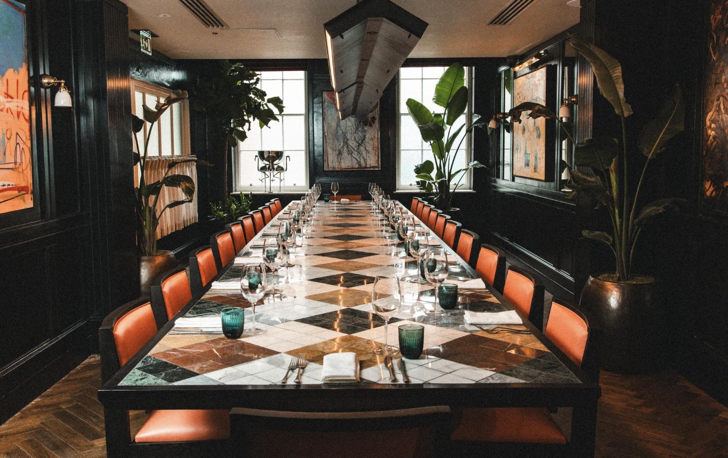A dining room with a long marble table, orange chairs, large windows, and potted plants at The Coal Shed - Brighton.