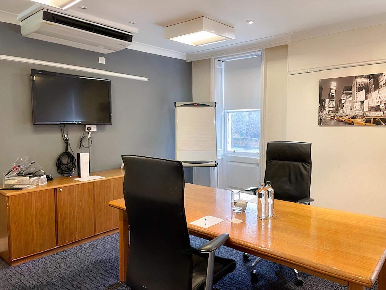 A hotel meeting room with a wooden conference table, wall-mounted TV, flip chart, and large windows at Mercure Thurrock Stifford Hall Hotel.