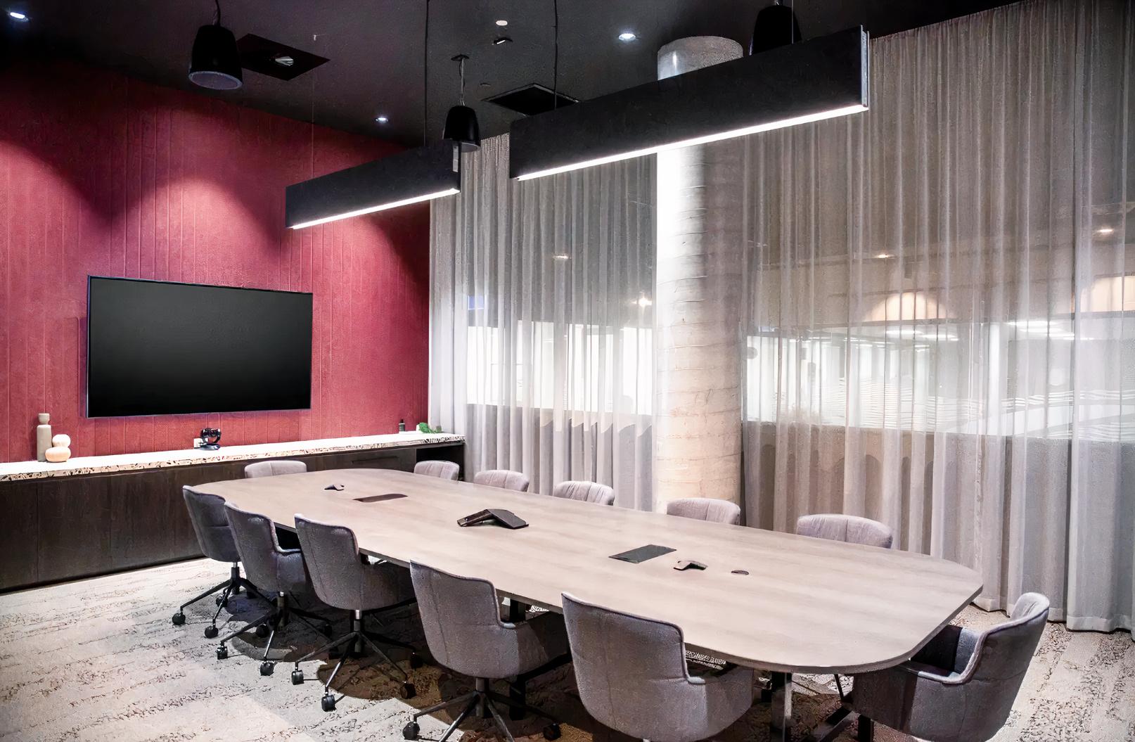 A meeting room with a long boardroom table, gray swivel chairs, wall-mounted screen, and sheer curtains at Waterman at Eastland.