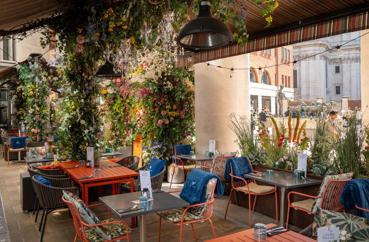 A restaurant terrace with floral arrangements, striped awnings, and colorful tables at The Happenstance – Paternoster Square.