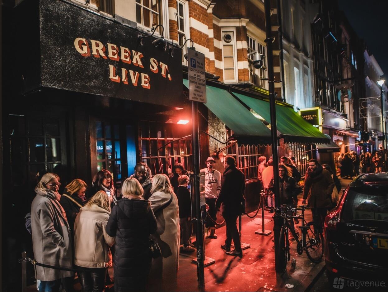 A pub exterior with a black sign reading 'Greek St. Live,' red window bars, and people gathered outside at Greek Street Live.