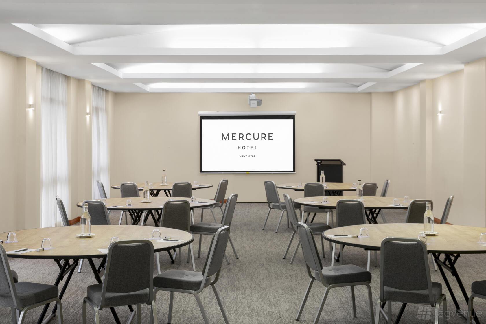 A meeting room with round tables, grey chairs, carpeted floor, and a projector screen at Mercure Newcastle.