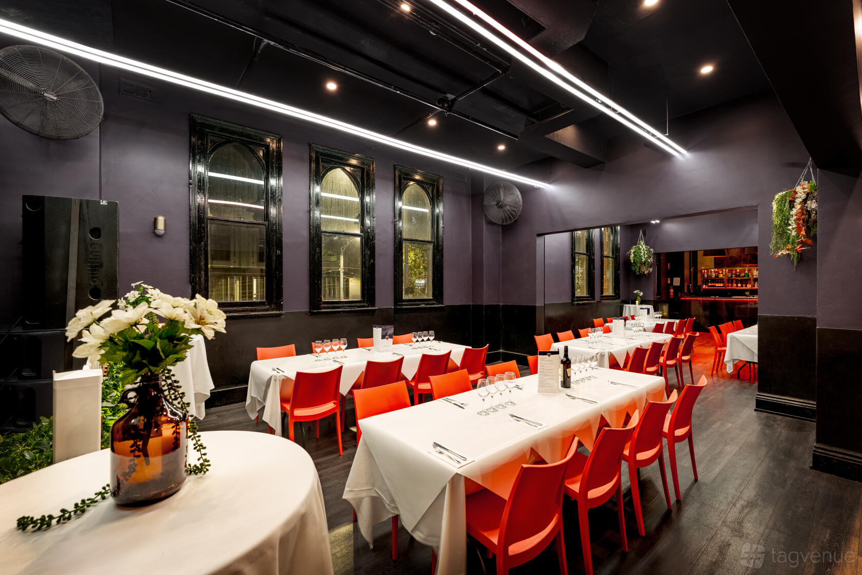An event space in a pub with arched windows, dark walls, red chairs, and white tablecloths at Royal Melbourne Hotel.