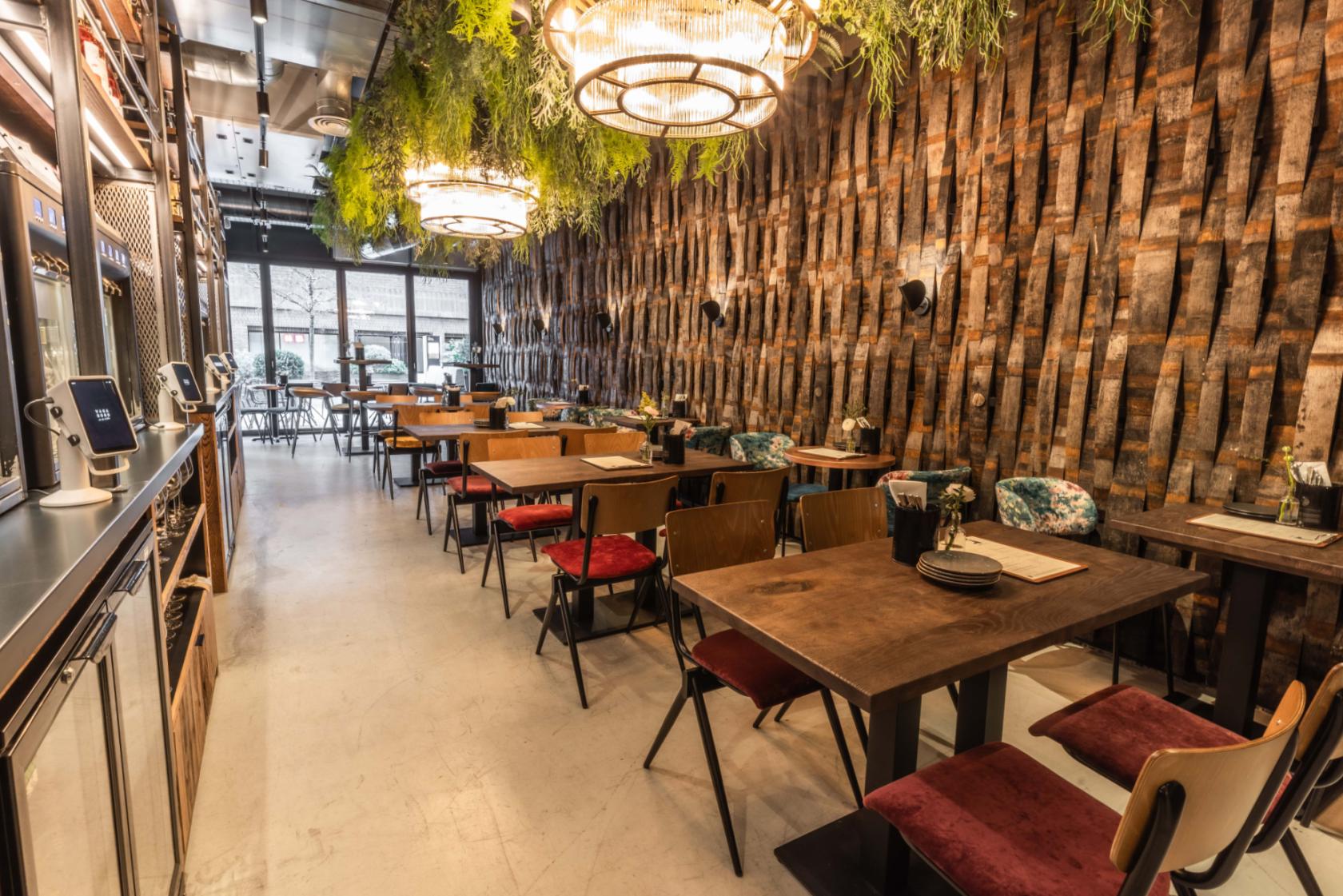 An event space in a bar with a wall of upright vintage barrels, hanging greenery, and wooden tables at Vagabond Battersea.