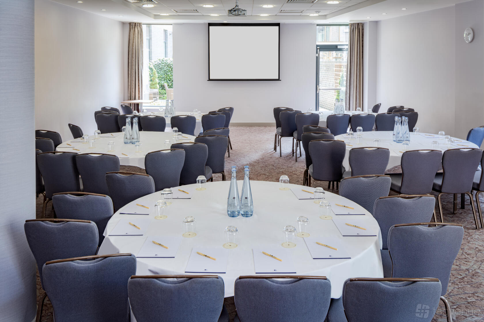 An event space with round tables covered in white linens, water bottles, and a projection screen at Doubletree by Hilton Angel Kings Cross.
