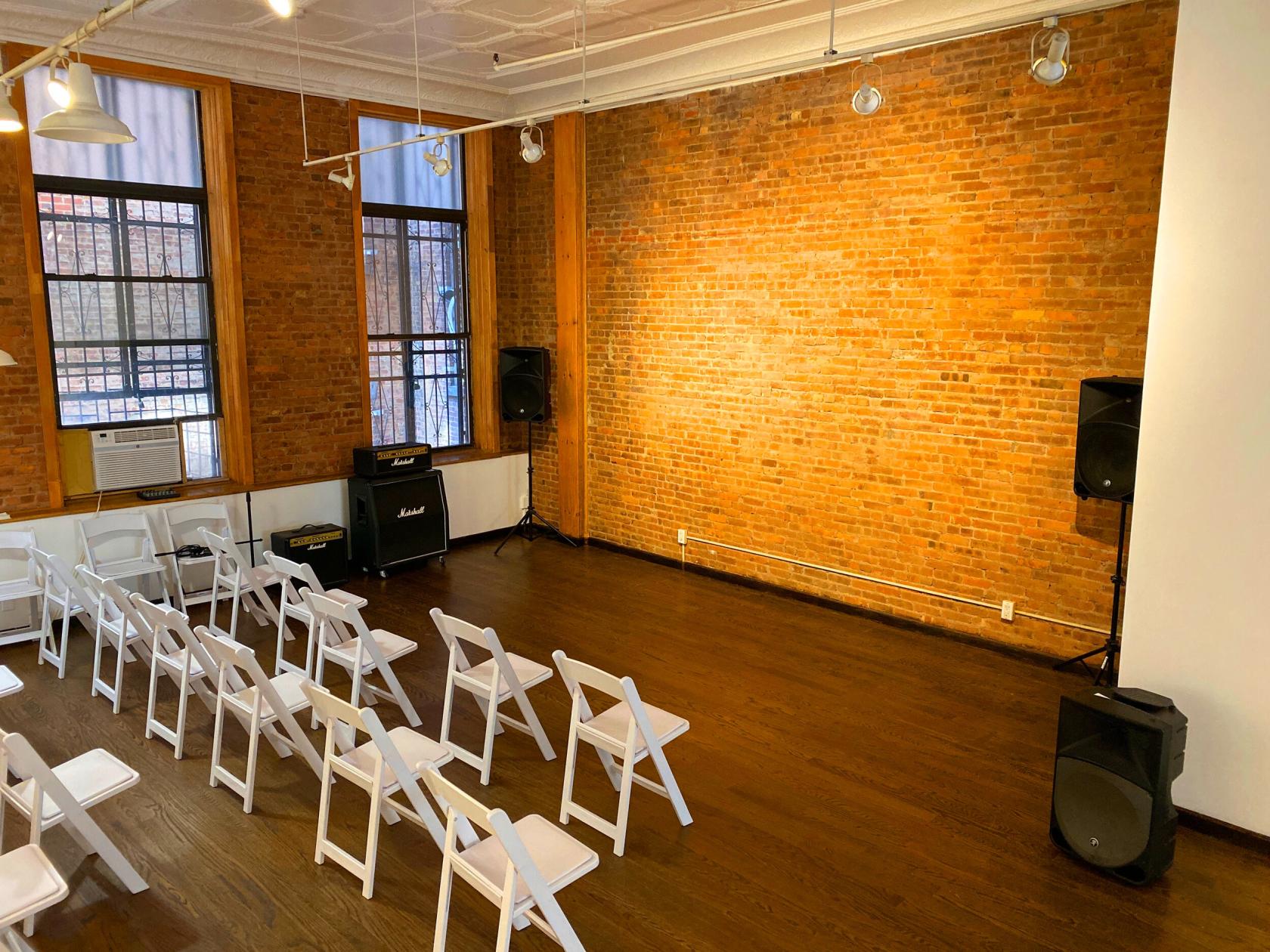 A loft with exposed brick walls, tall windows, and rows of white folding chairs facing a small stage at LOFT393.