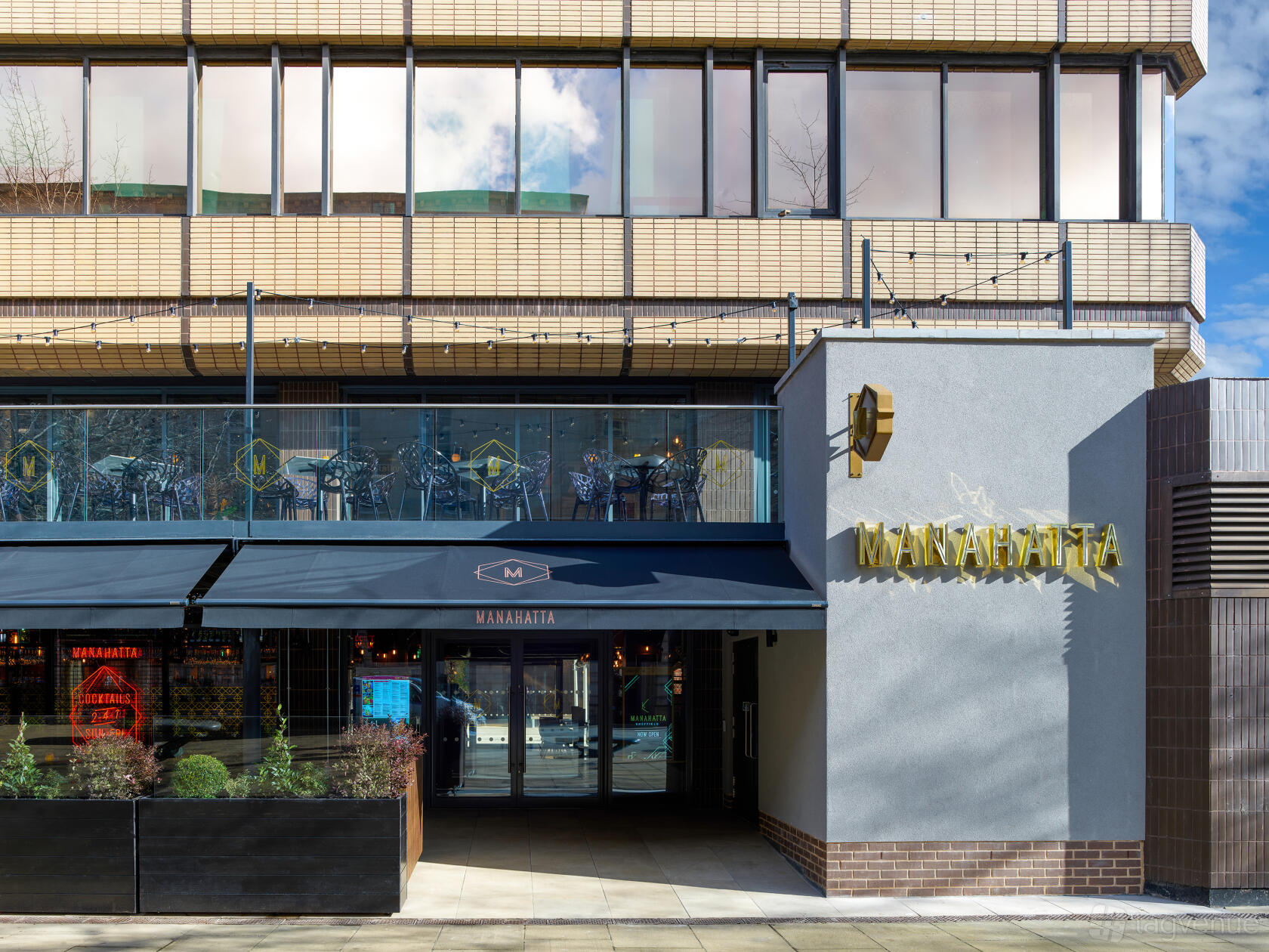 A pub with a glass-walled upper level, exterior awning, and gold signage at Manahatta Sheffield.