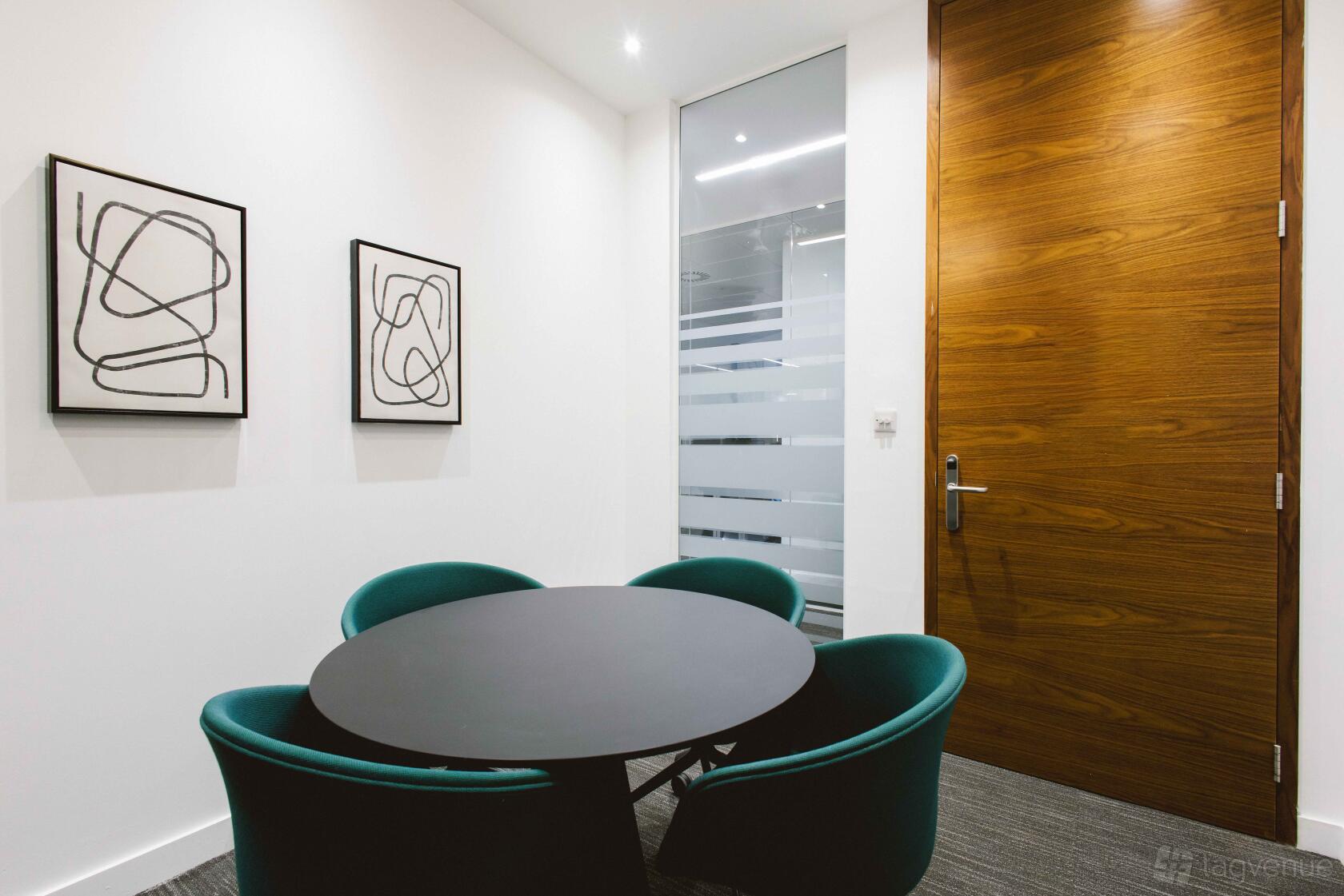 A boardroom with a round black table, green chairs, abstract wall art, and a wooden door at Orega Gracechurch Street.