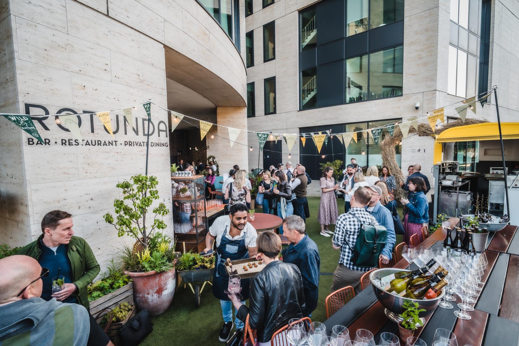 A restaurant terrace with outdoor seating, artificial grass, bunting décor, and guests mingling at Rotunda Bar and Restaurant.