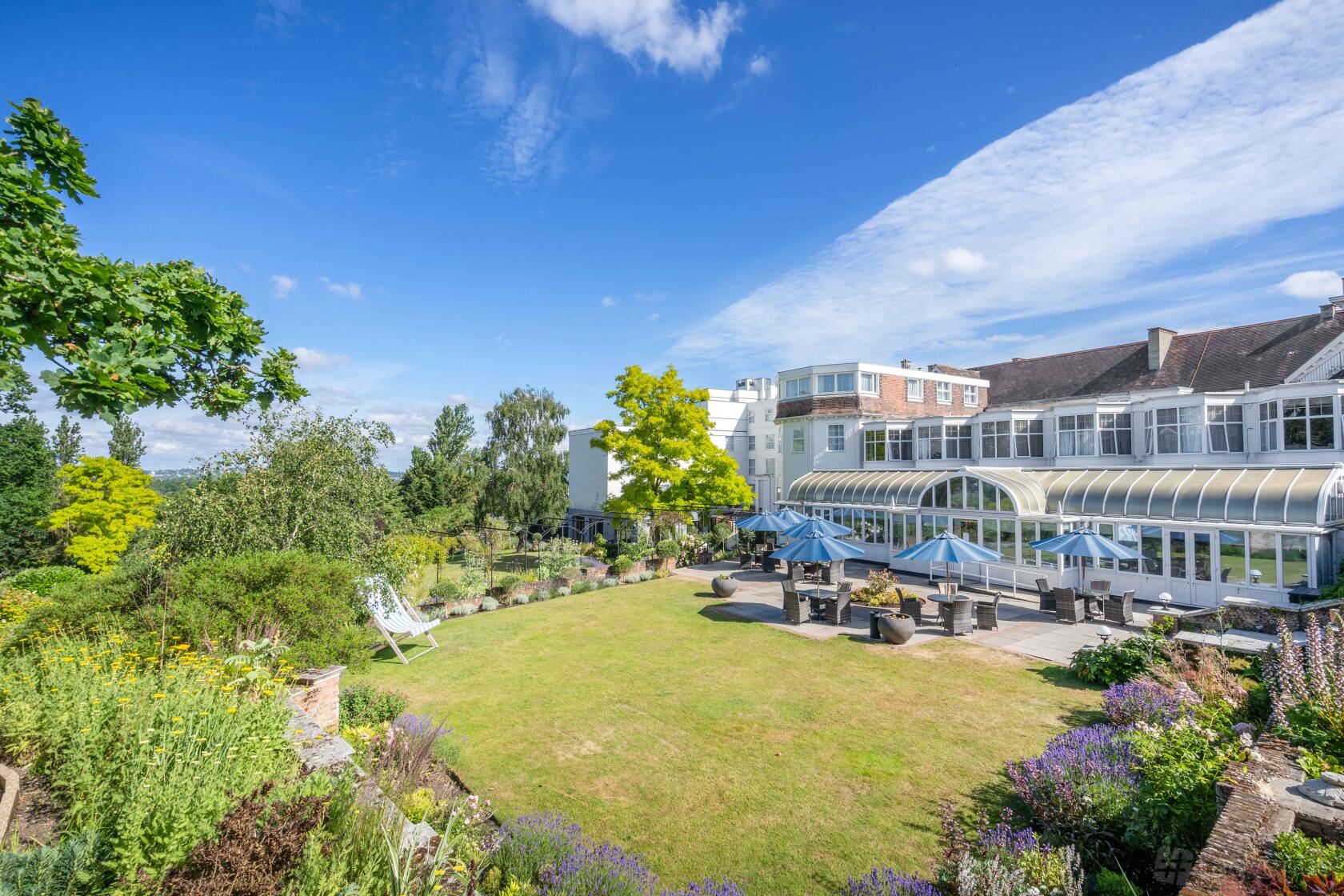 A hotel with landscaped gardens, outdoor seating areas with umbrellas, and large windows at The Bromley Court Hotel.