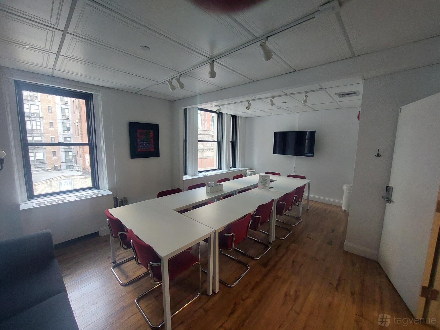 A meeting room with two large windows, white tables arranged in a U-shape, red chairs, and a wall-mounted screen at Sage Workspace.