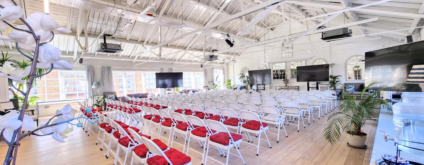 An event venue with white exposed beams, folding chairs with red cushions, and large windows at Lumiere London Old Street.
