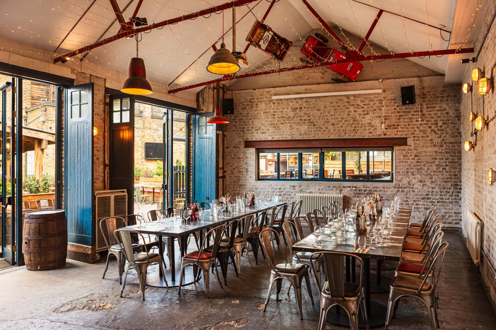A function room with exposed brick walls, long banquet tables, industrial chairs, and open double doors at The Half Moon.